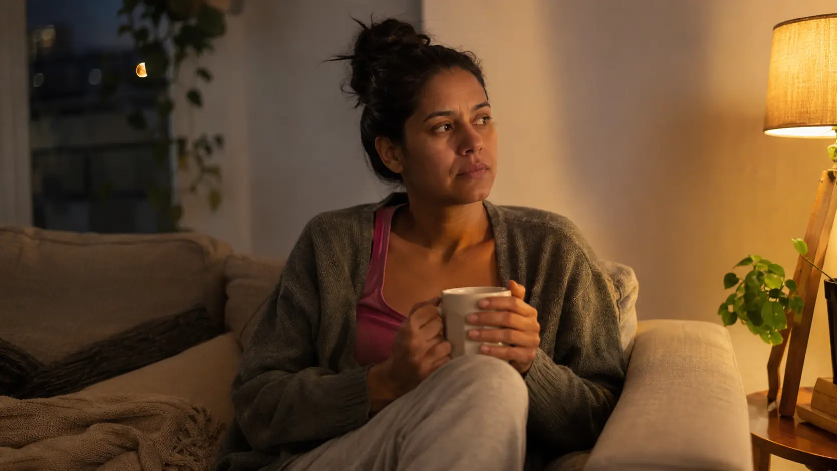 Mulher sentada sozinha em casa à noite, com expressão pensativa segurando uma caneca