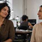 Duas colegas de trabalho num escritório, uma fazendo um comentário com meio sorriso e a outra ouvindo com postura tranquila