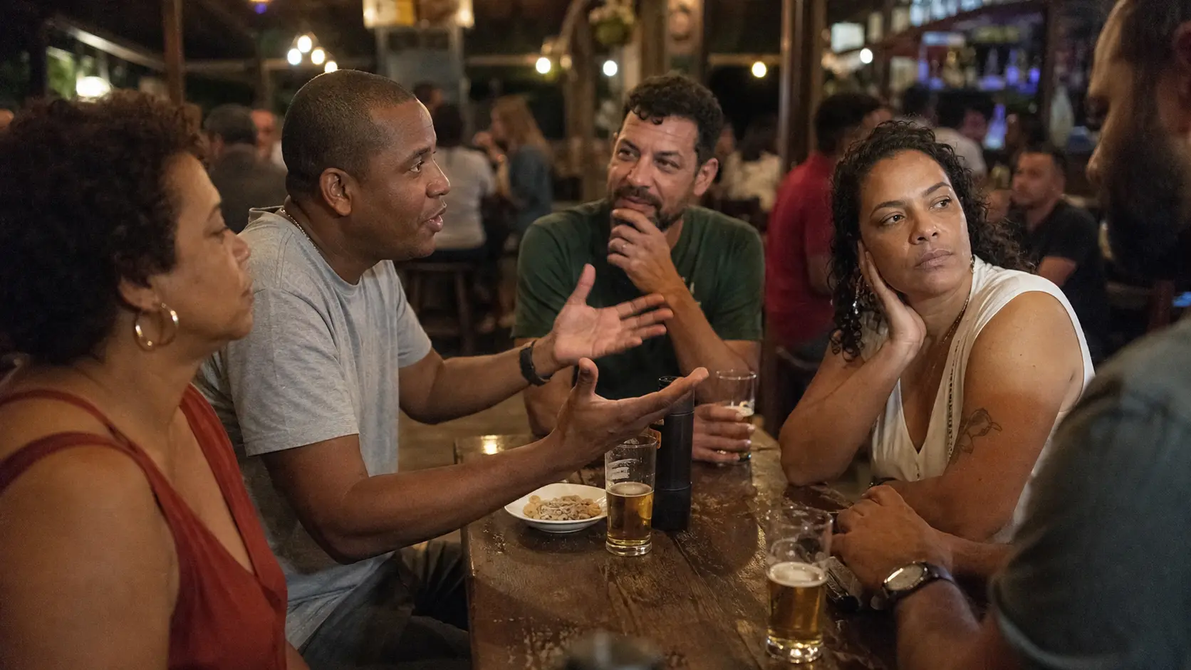Grupo de amigos numa mesa de bar, uma pessoa discutindo com o grupo enquanto outra se desliga da discussão